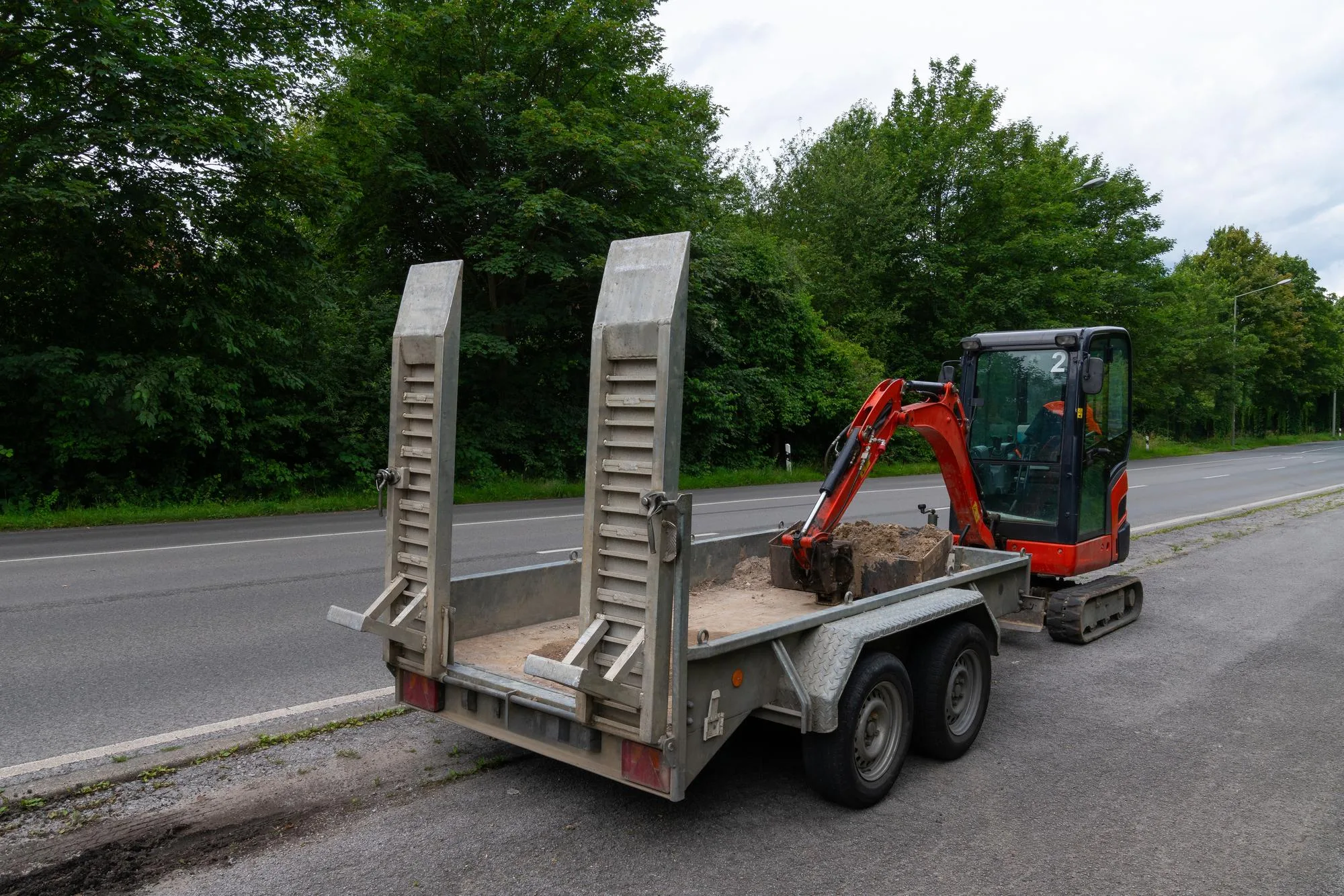 Mini excavator loaded onto a dual-axle trailer with raised loading ramps, parked on the roadside beside a dense green wooded area.