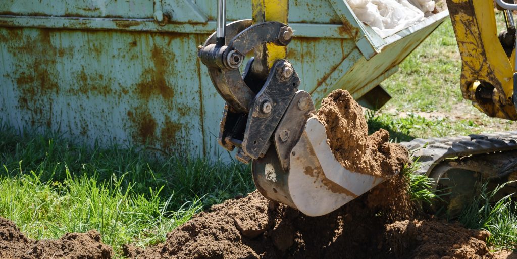 Mini excavator digs a narrow trench in soil for laying communications close-up. Mini bucket of crawler machine. Earthworks with heavy special equipment. Digging machinery works at construction site