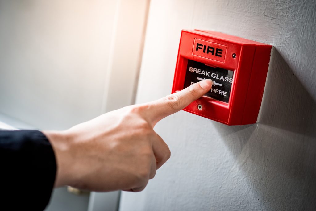Male hand pointing at red fire alarm switch on concrete wall in office building. Industrial fire warning system equipment for emergency.