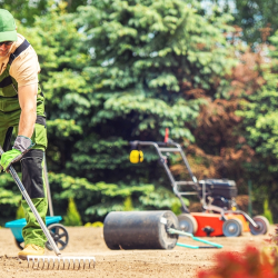 A landscaper wearing protective gear rakes soil in a garden area on a sunny day, with lawn equipment and greenery in the background.