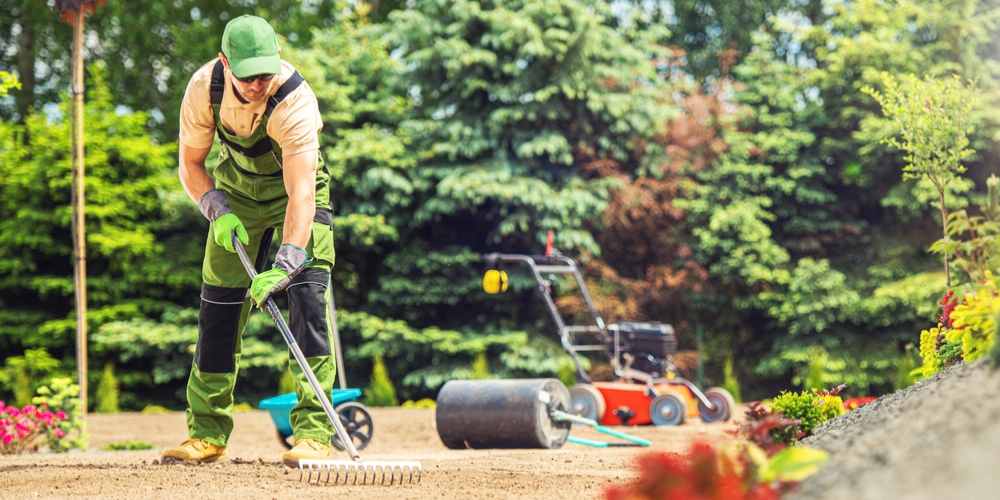 Blog-Image-Crop-1000x500 (10) A landscaper wearing protective gear rakes soil in a garden area on a sunny day, with lawn equipment and greenery in the background.