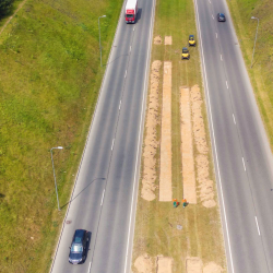 Aerial view of highway median landscaping work in progress, with workers and equipment installing sod between divided roadways as vehicles pass on both sides.
