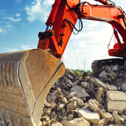 A large orange excavator sits on a pile of broken concrete and rubble at a construction or demolition site under a partly cloudy blue sky.