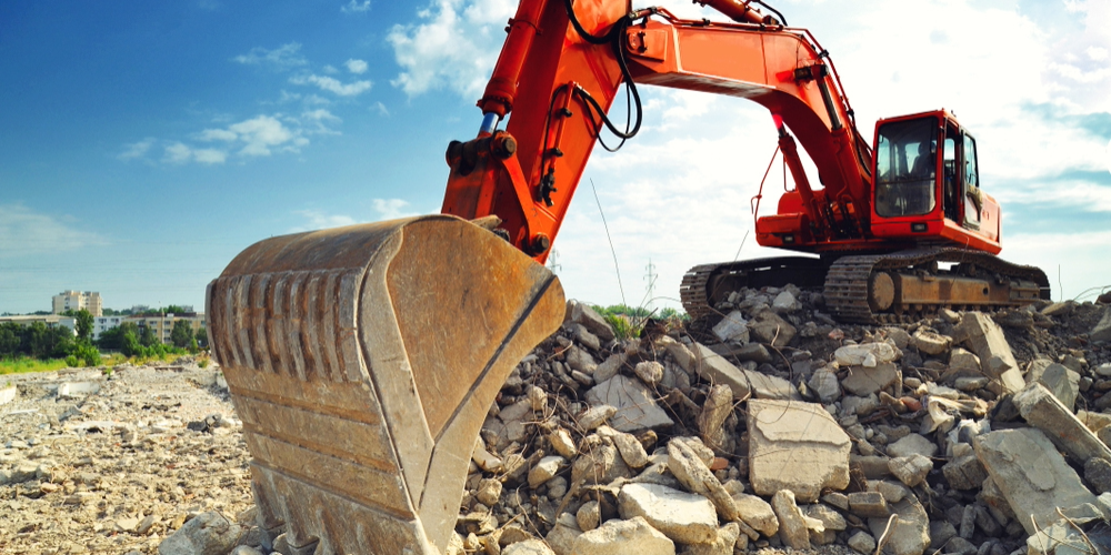 Blog-Image-Crop-1000x500 (9) A large orange excavator sits on a pile of broken concrete and rubble at a construction or demolition site under a partly cloudy blue sky.