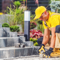 Professional landscaper kneeling to install stone pavers along a landscaped walkway.