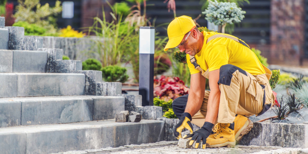 Professional landscaper kneeling to install stone pavers along a landscaped walkway.