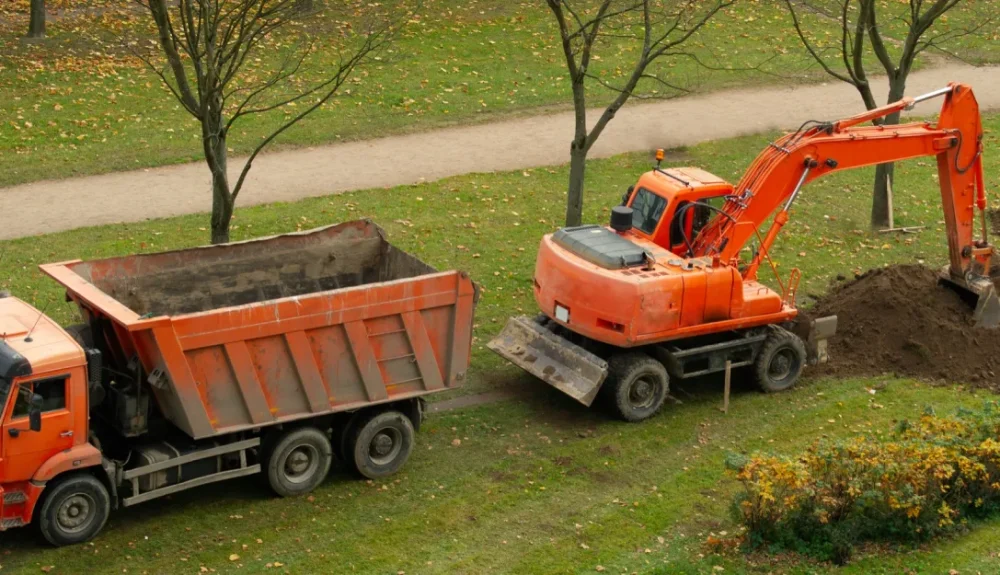 Orange excavator digging soil beside an orange dump truck at a landscaping and earthmoving site in a grassy park.
