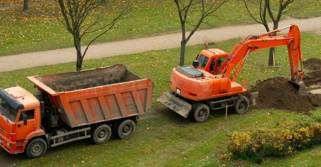 Orange excavator digging soil beside an orange dump truck at a landscaping and earthmoving site in a grassy park. Orange excavator digging soil beside an orange dump truck at a landscaping and earthmoving site in a grassy park.