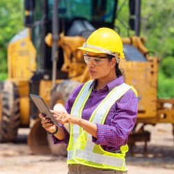A female construction worker wearing a yellow hard hat and safety vest stands on a worksite, using a tablet. Heavy machinery is visible in the background, and the worker appears focused on the task at hand.