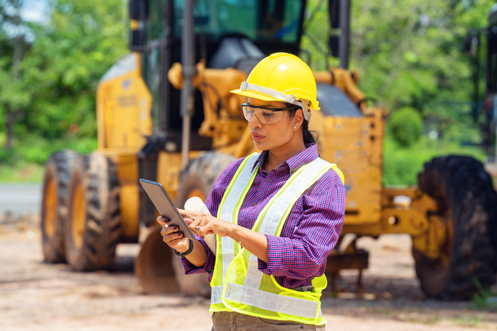 A female construction worker wearing a yellow hard hat and safety vest stands on a worksite, using a tablet. Heavy machinery is visible in the background, and the worker appears focused on the task at hand.