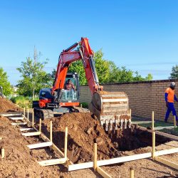 A construction worker operates an orange excavator at a work site, digging soil for a foundation, while another worker stands nearby. The site is framed with wooden structures, preparing for construction, and is surrounded by greenery and a brick wall under a clear blue sky.