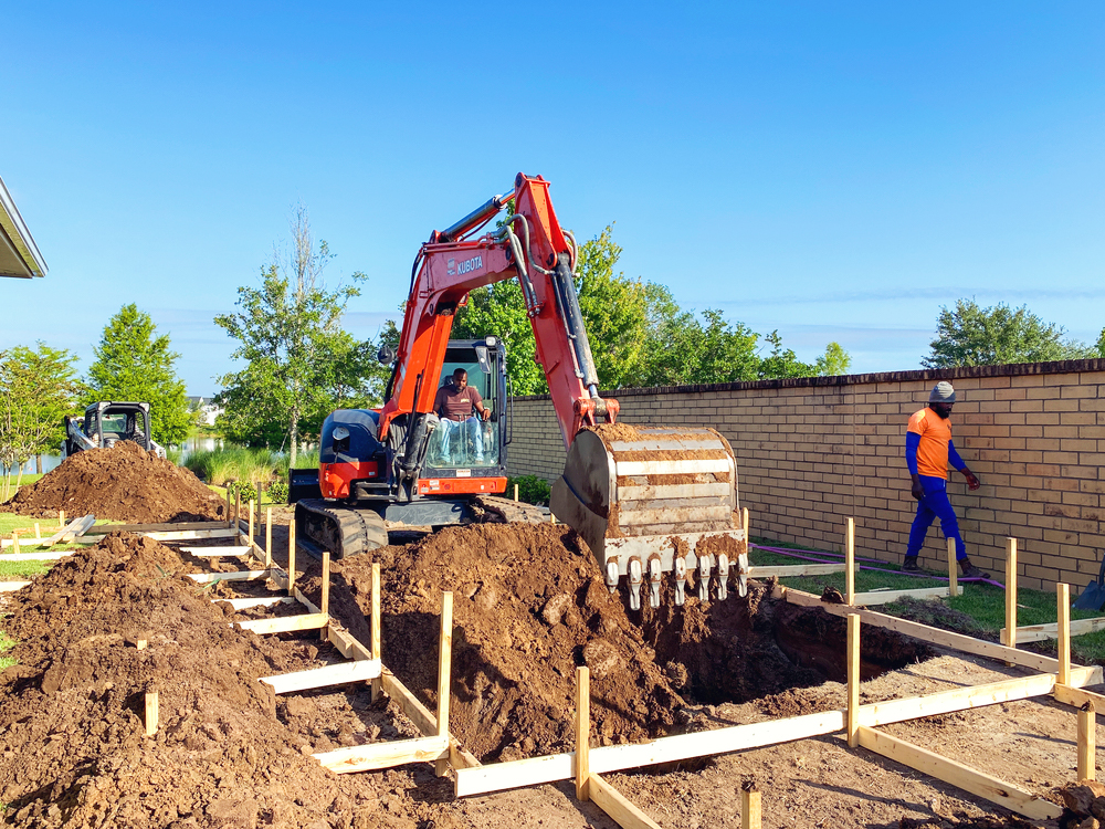 A construction worker operates an orange excavator at a work site, digging soil for a foundation, while another worker stands nearby. The site is framed with wooden structures, preparing for construction, and is surrounded by greenery and a brick wall under a clear blue sky.