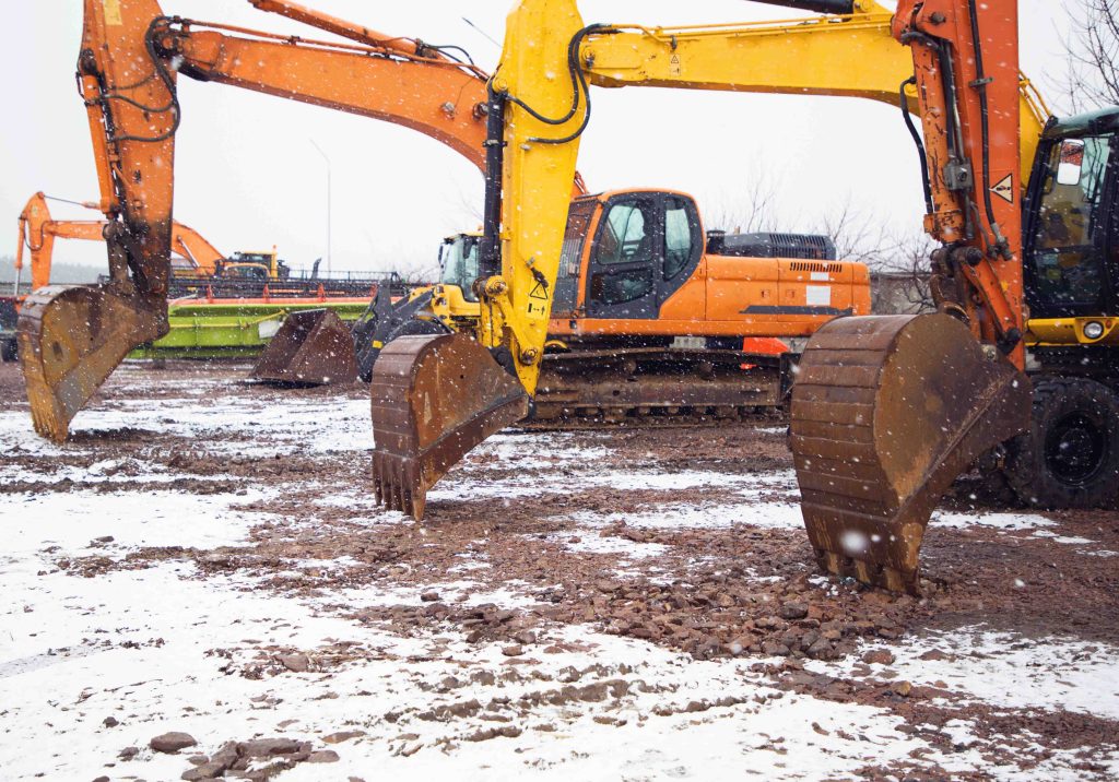 large orange and yellow used crawler excavators standing side by side at a construction site. Construction equipment sits idle on a snow-covered site, highlighting the challenges of winter construction delays and cold-weather project risks.