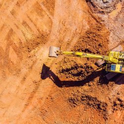 Top-down view of a yellow excavator digging into red earth at a construction site, with visible tracks and soil patterns around the machine.