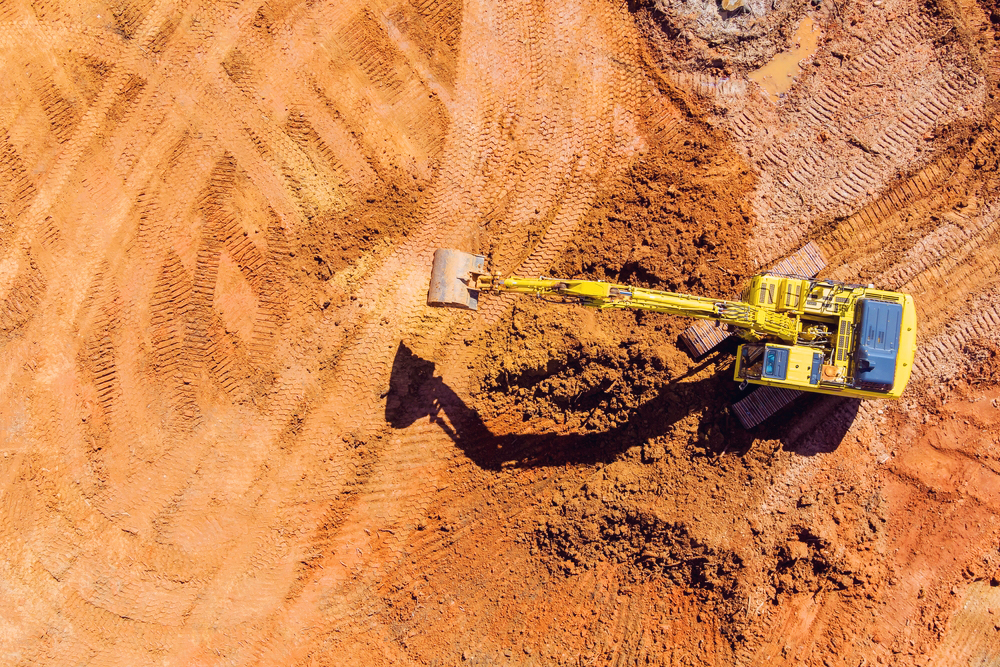 Top-down view of a yellow excavator digging into red earth at a construction site, with visible tracks and soil patterns around the machine.