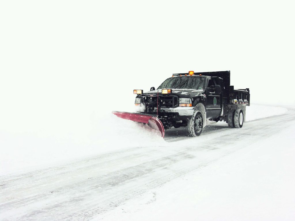 shutterstock_712240 A snowplow truck clearing a snow-covered road on a winter day, with heavy snow visible on the ground and surrounding area.