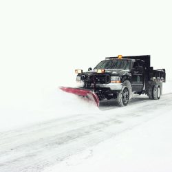 A snowplow truck clearing a snow-covered road on a winter day, with heavy snow visible on the ground and surrounding area.